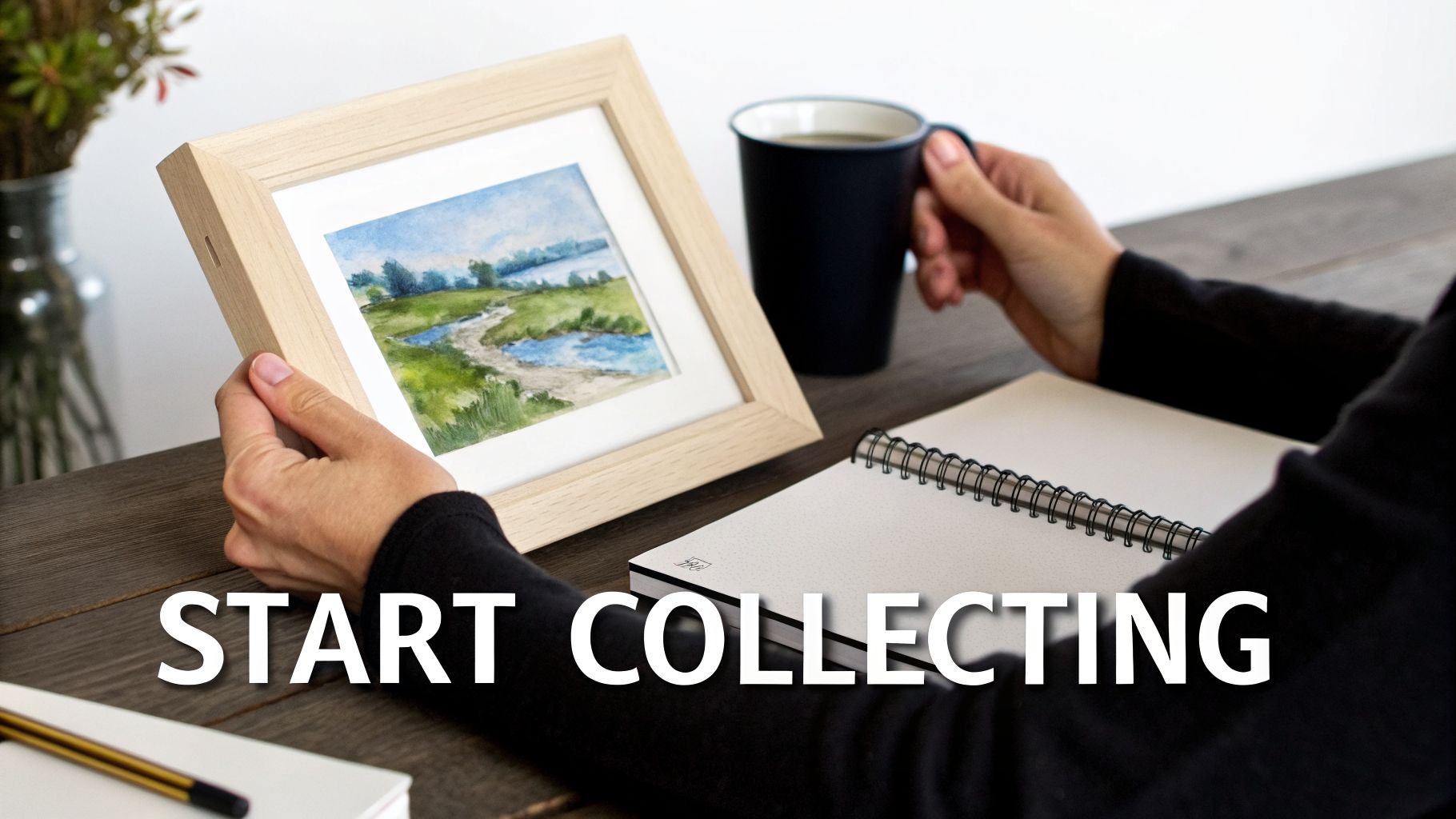 A person holding a framed watercolor painting of a landscape, next to a coffee mug and notebook on a table.