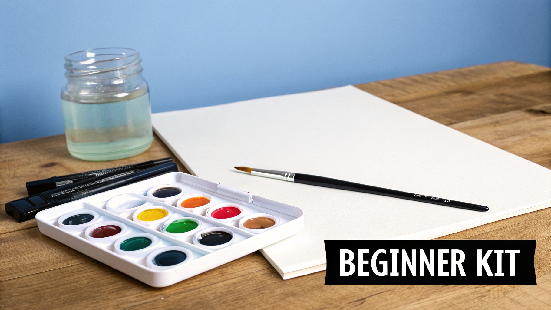 A beginner's watercolor painting kit with paints, brushes, water, and paper on a wooden table.