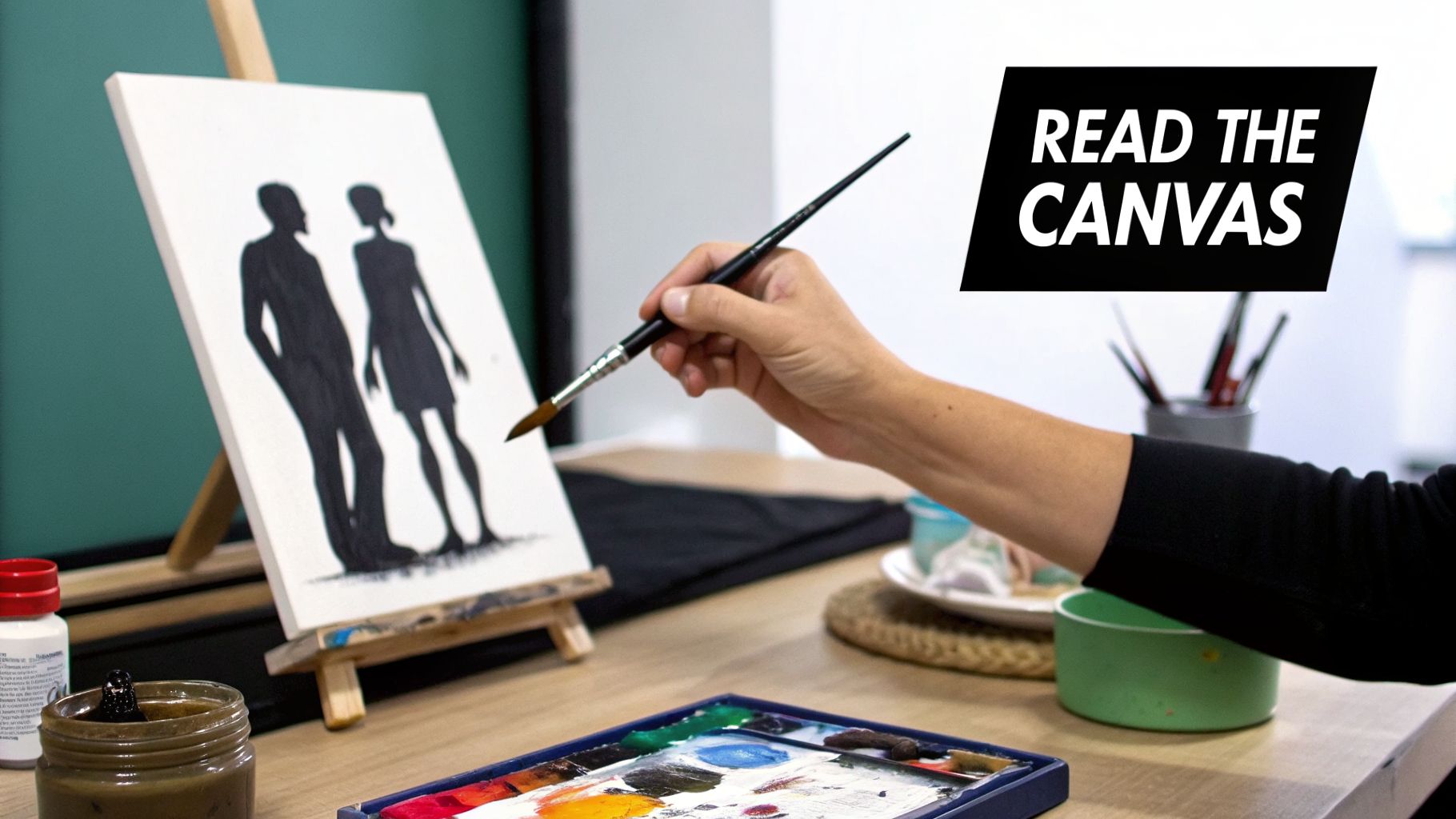 A person's hand painting black silhouettes of two people on a white canvas on an easel.